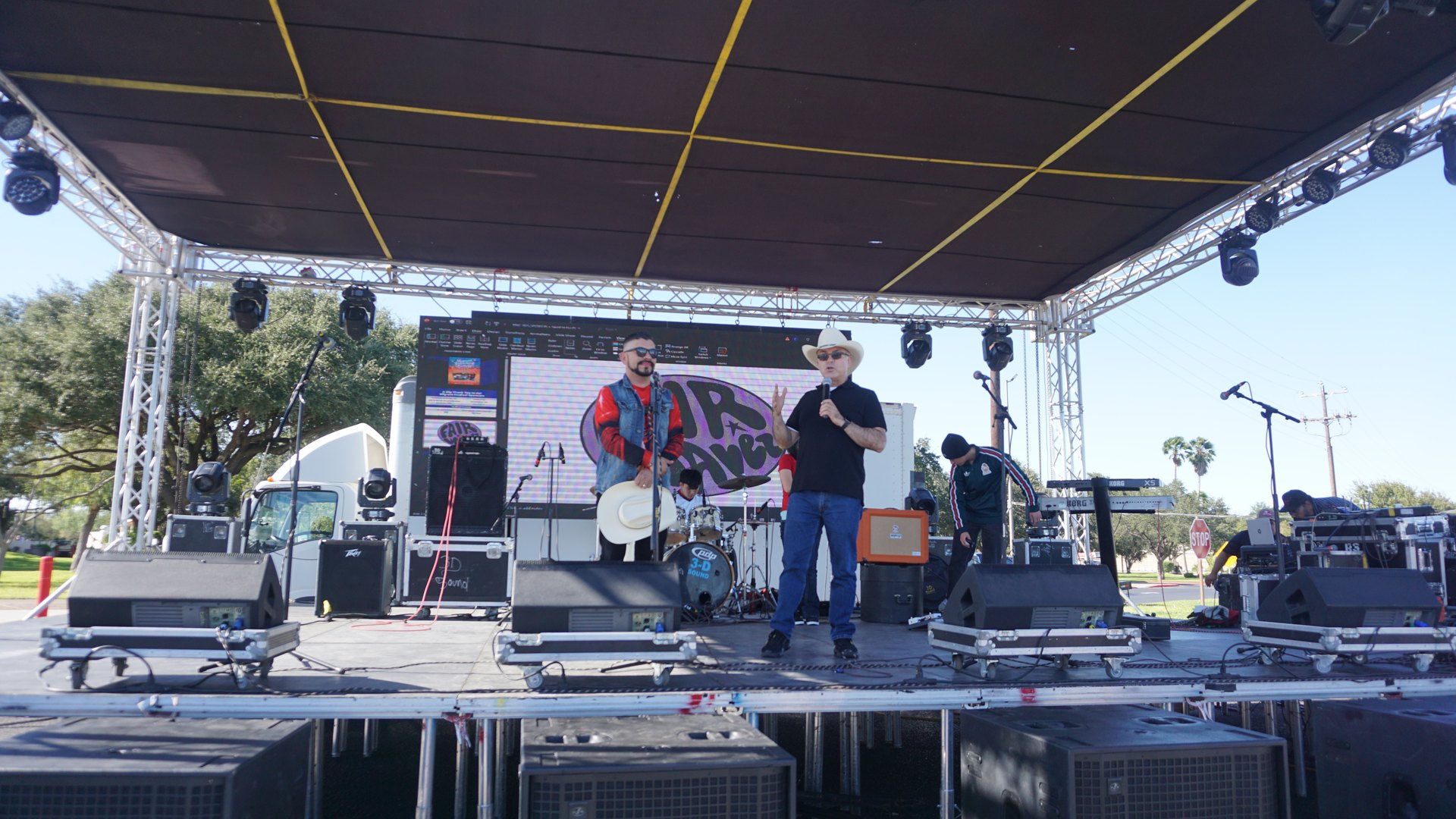 Two presenters at microphones on the outdoor festival stage, with musicians and a drum kit behind them.