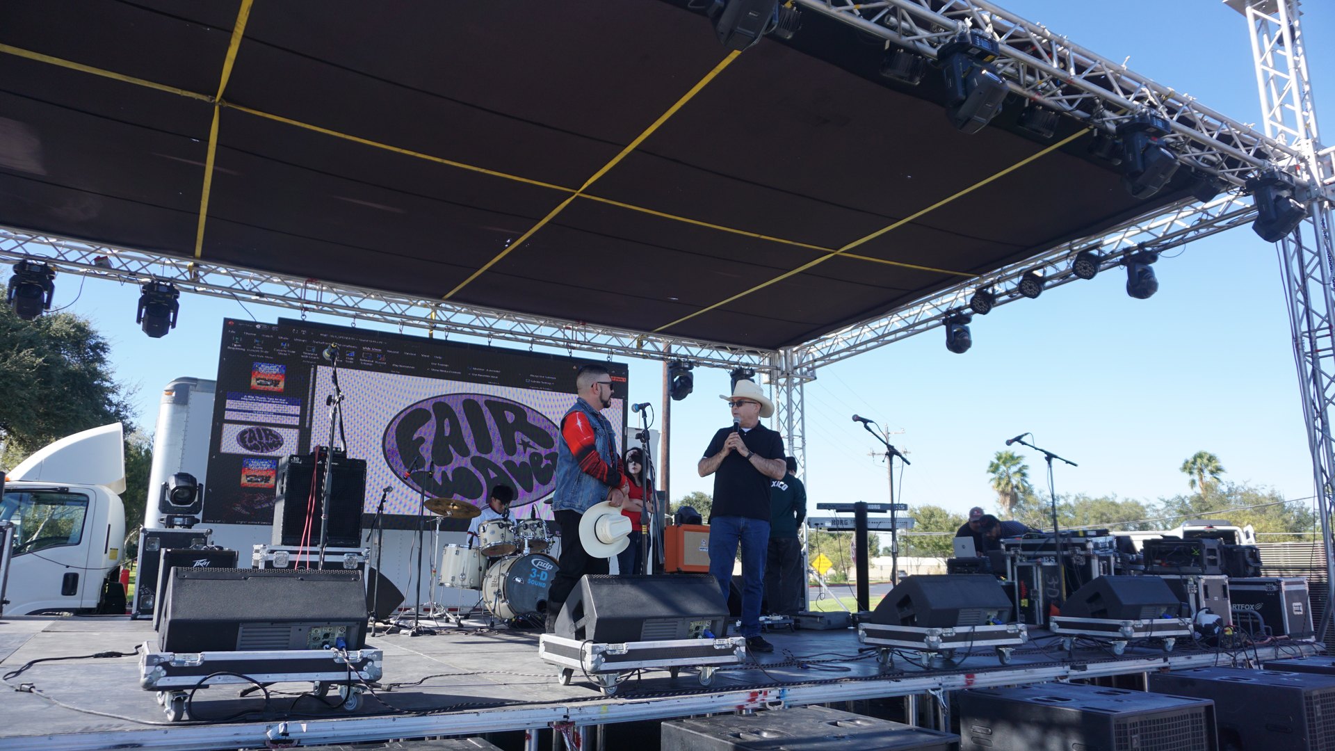Presenters stand at the center of the covered festival stage, surrounded by speakers and musical equipment.