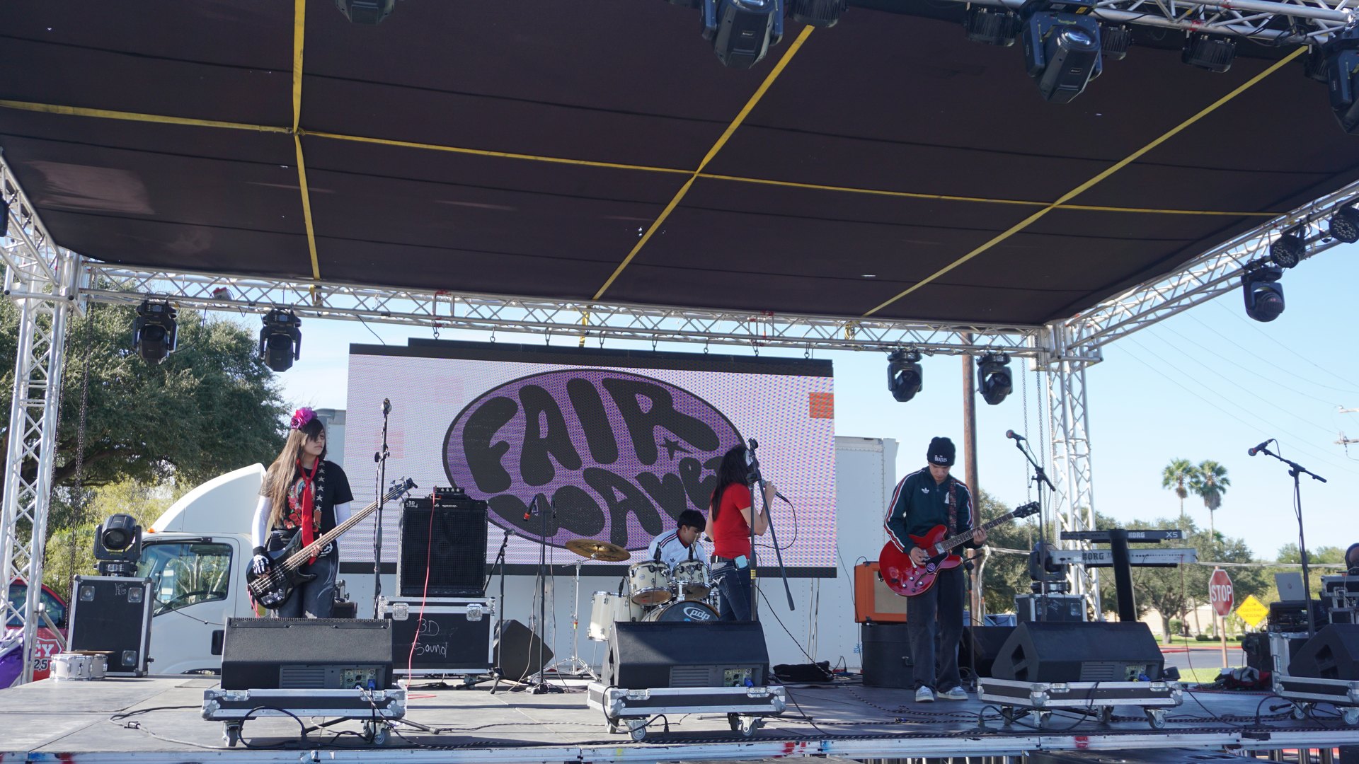 A band performs on the festival stage with guitars, microphones, a drum kit and stage speakers.