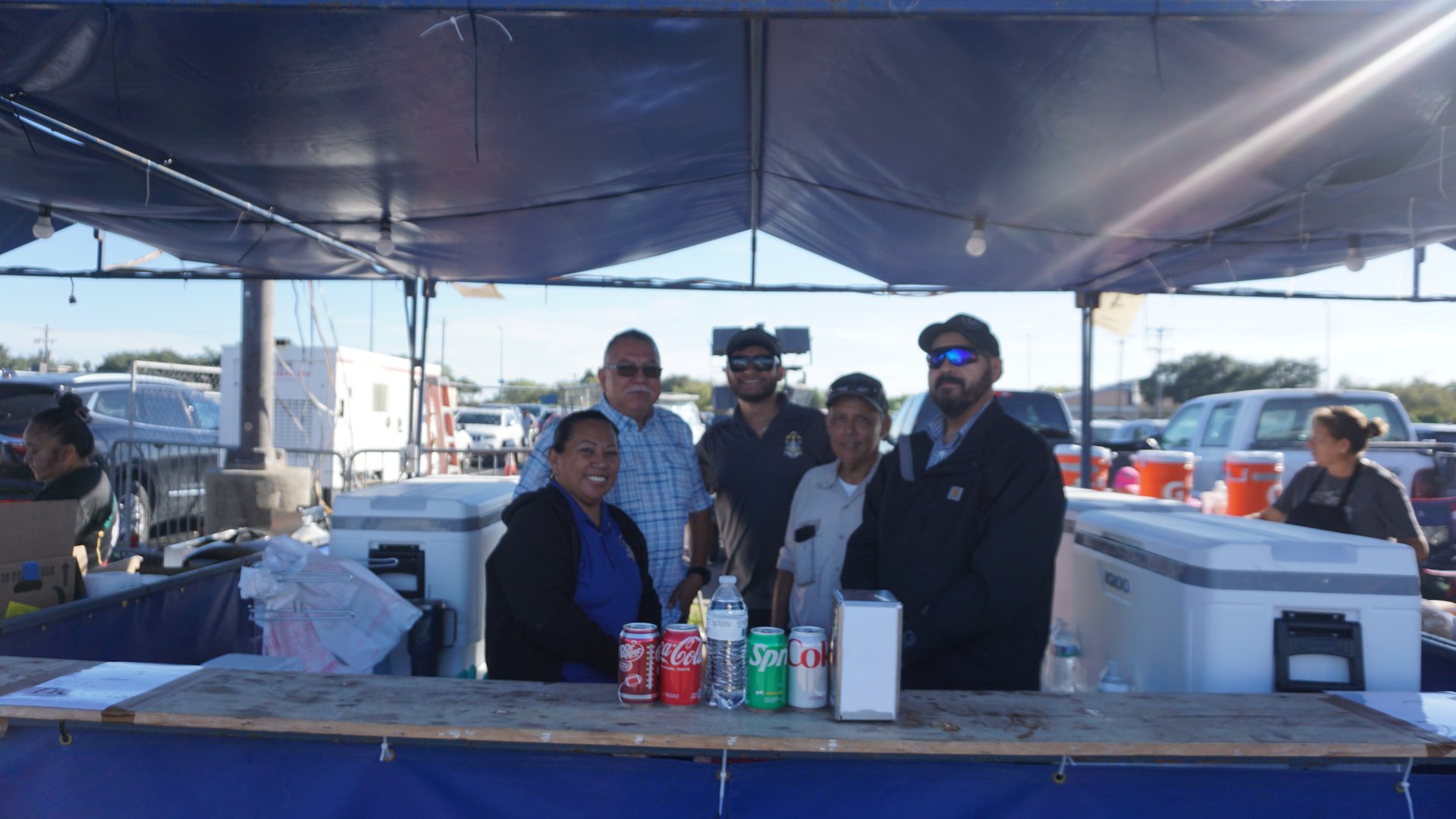 Festival booth staff pose behind a concession counter stocked with drinks and coolers.