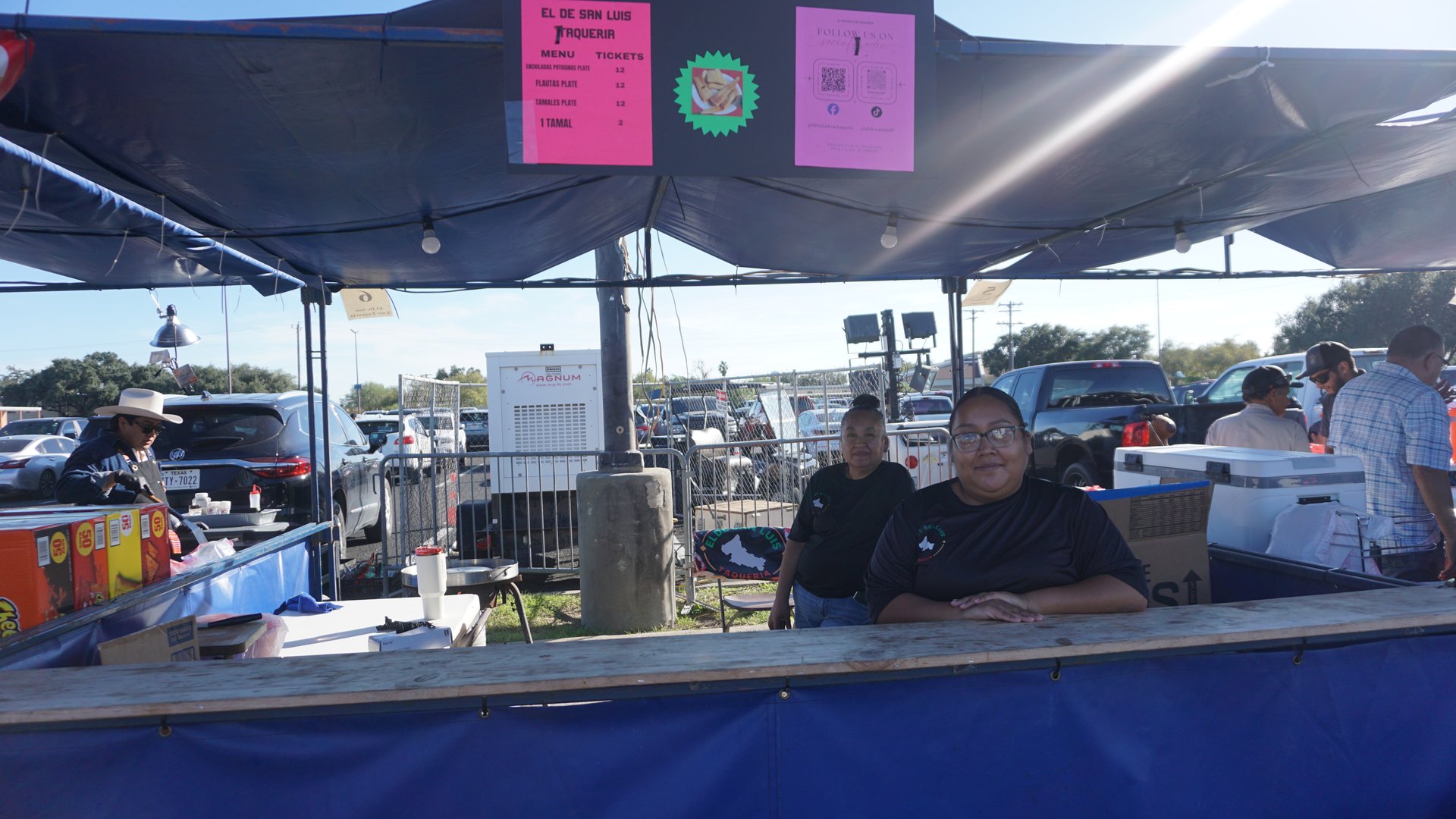 Two booth attendants sit behind a blue concession counter beneath bright pink menu signs.