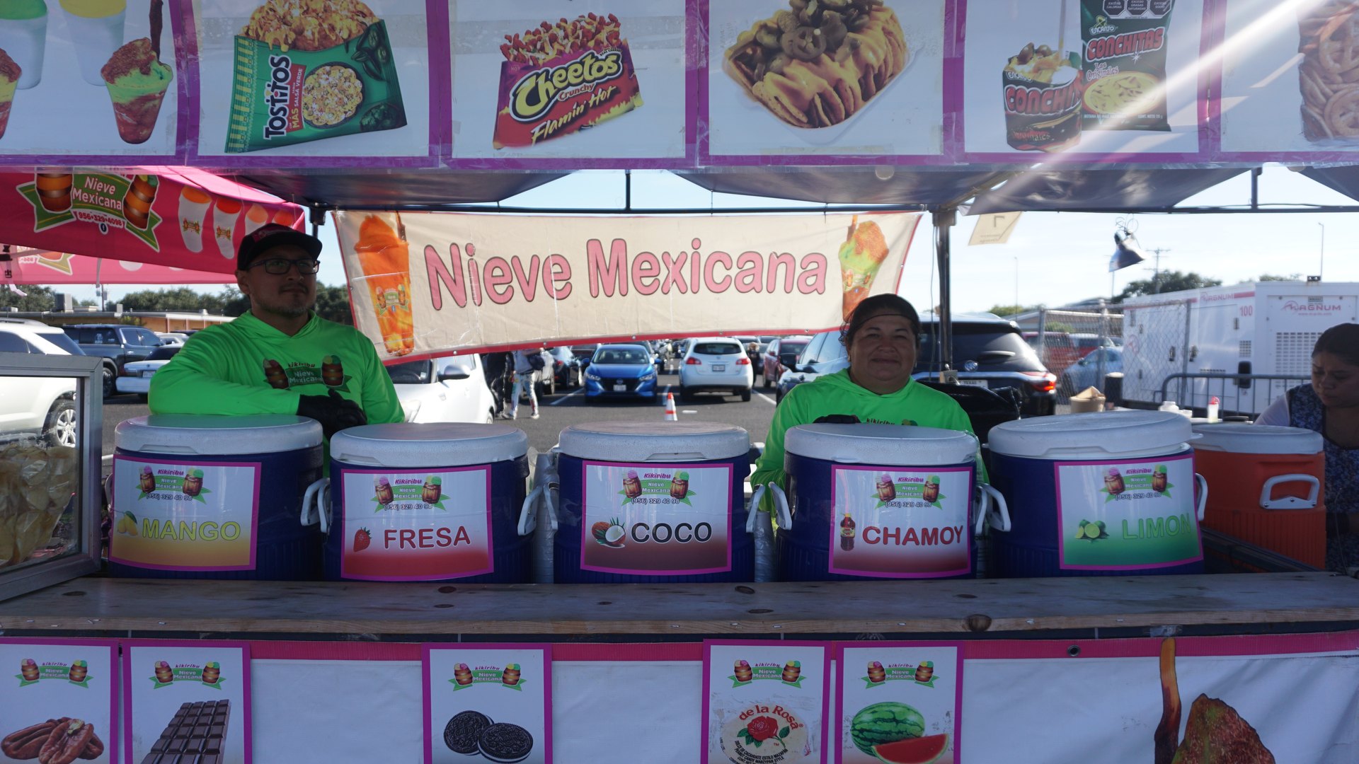 Two vendors in green shirts stand behind a Nieve Mexicana frozen treat counter with labeled tubs and a colorful menu.
