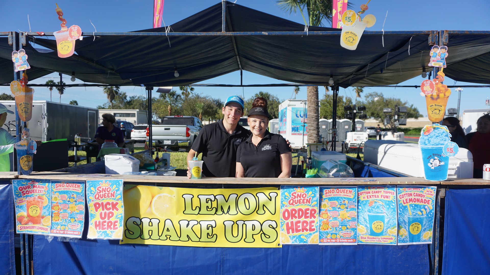 Two smiling vendors pose at a colorful Lemon Shake Ups stand surrounded by lemonade and frozen drink menus.