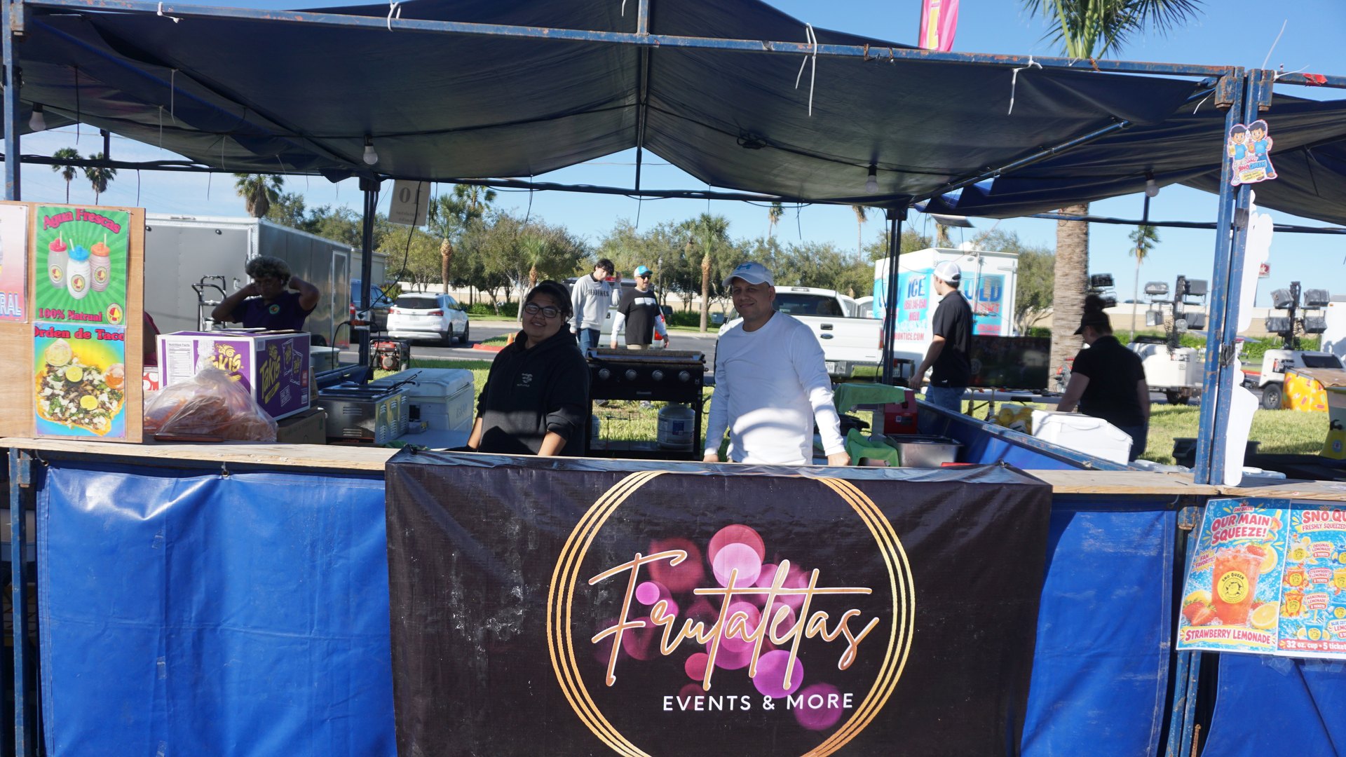 Three booth attendants stand behind a blue concession counter beneath a Fiestitas Events and More banner.