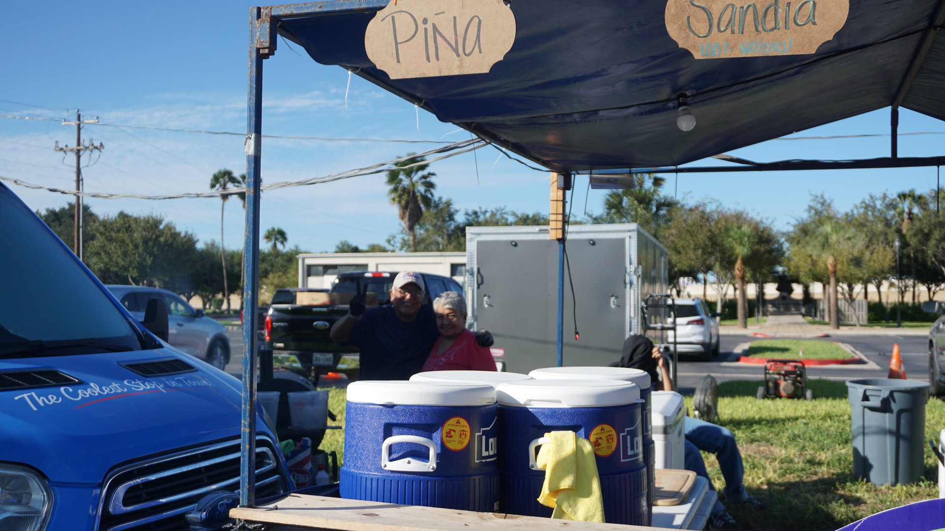 A vendor sits behind drink coolers beneath pineapple and watermelon signs at an outdoor festival booth.