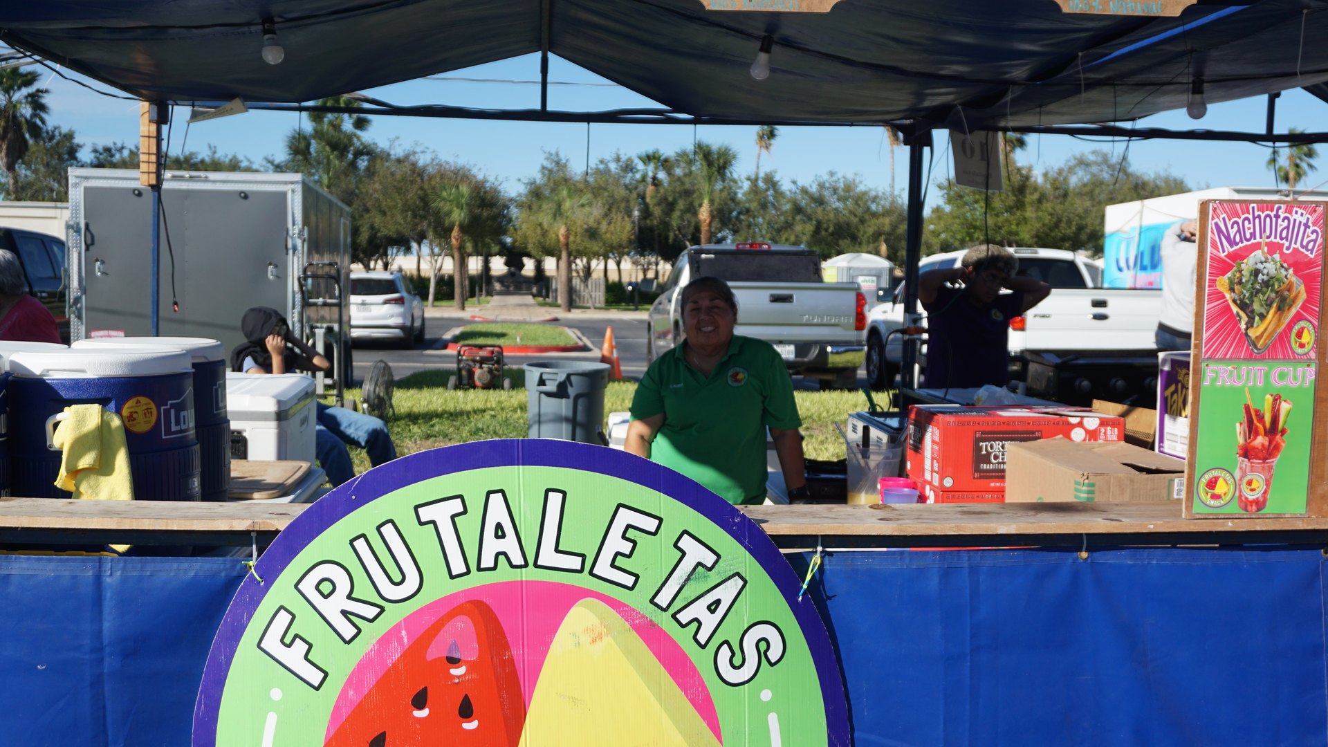 A vendor stands at a blue festival booth decorated with a large Frutaletas watermelon sign.