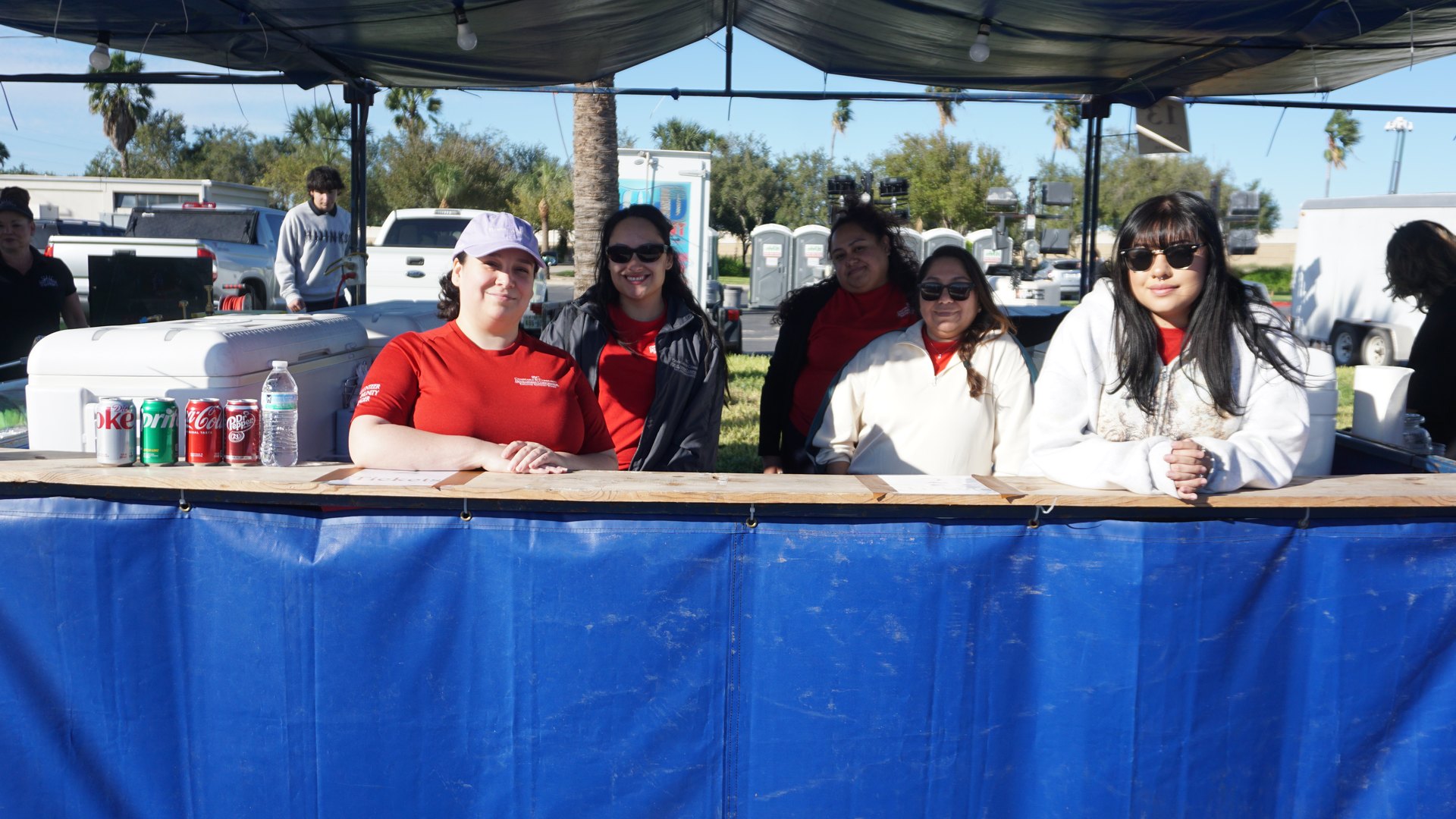 Five smiling booth staff gather behind a blue concession counter with drinks and coolers at the festival.