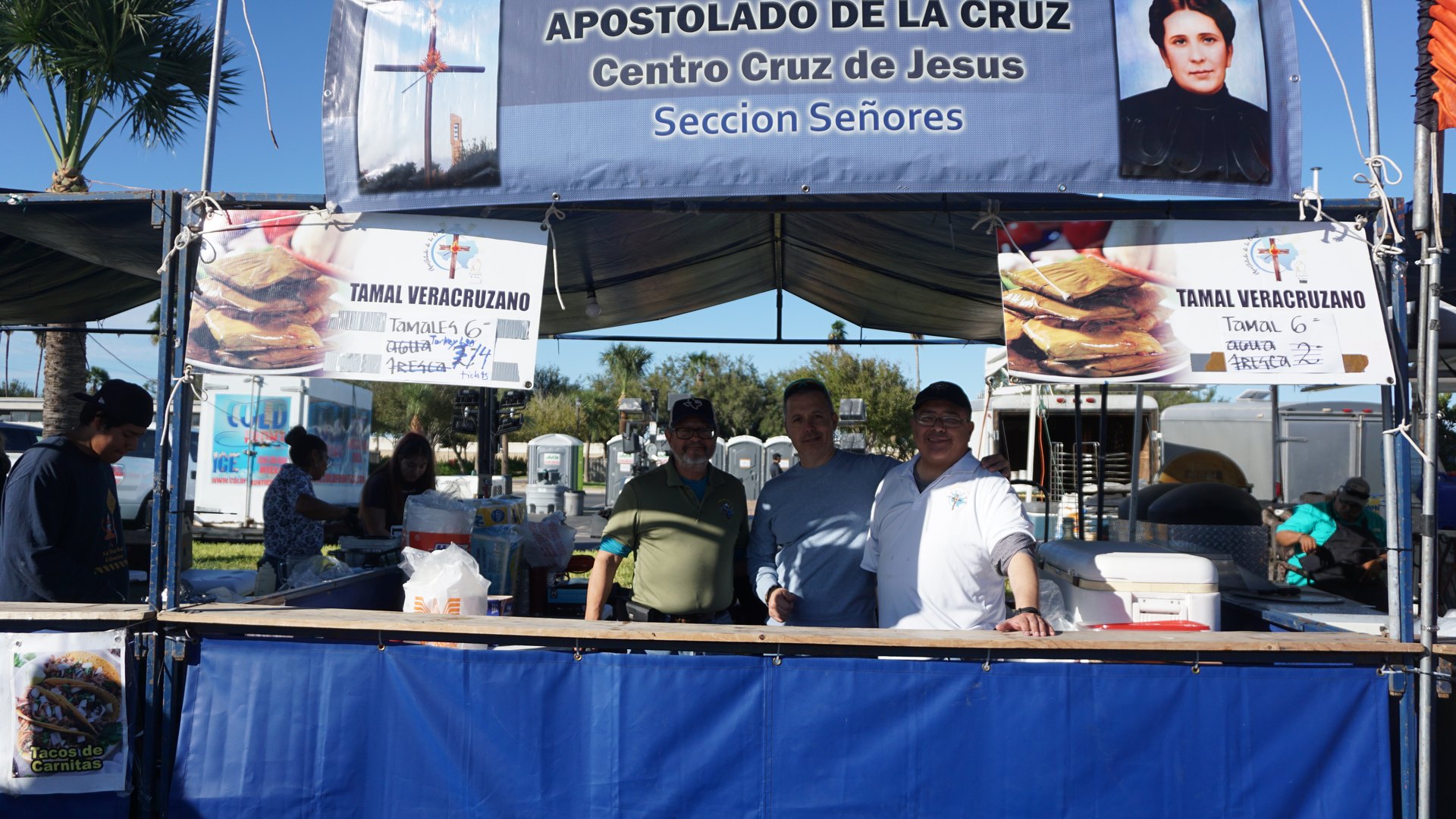 Three booth attendants pose at a tamale stand beneath a large Apostolado de la Cruz banner.