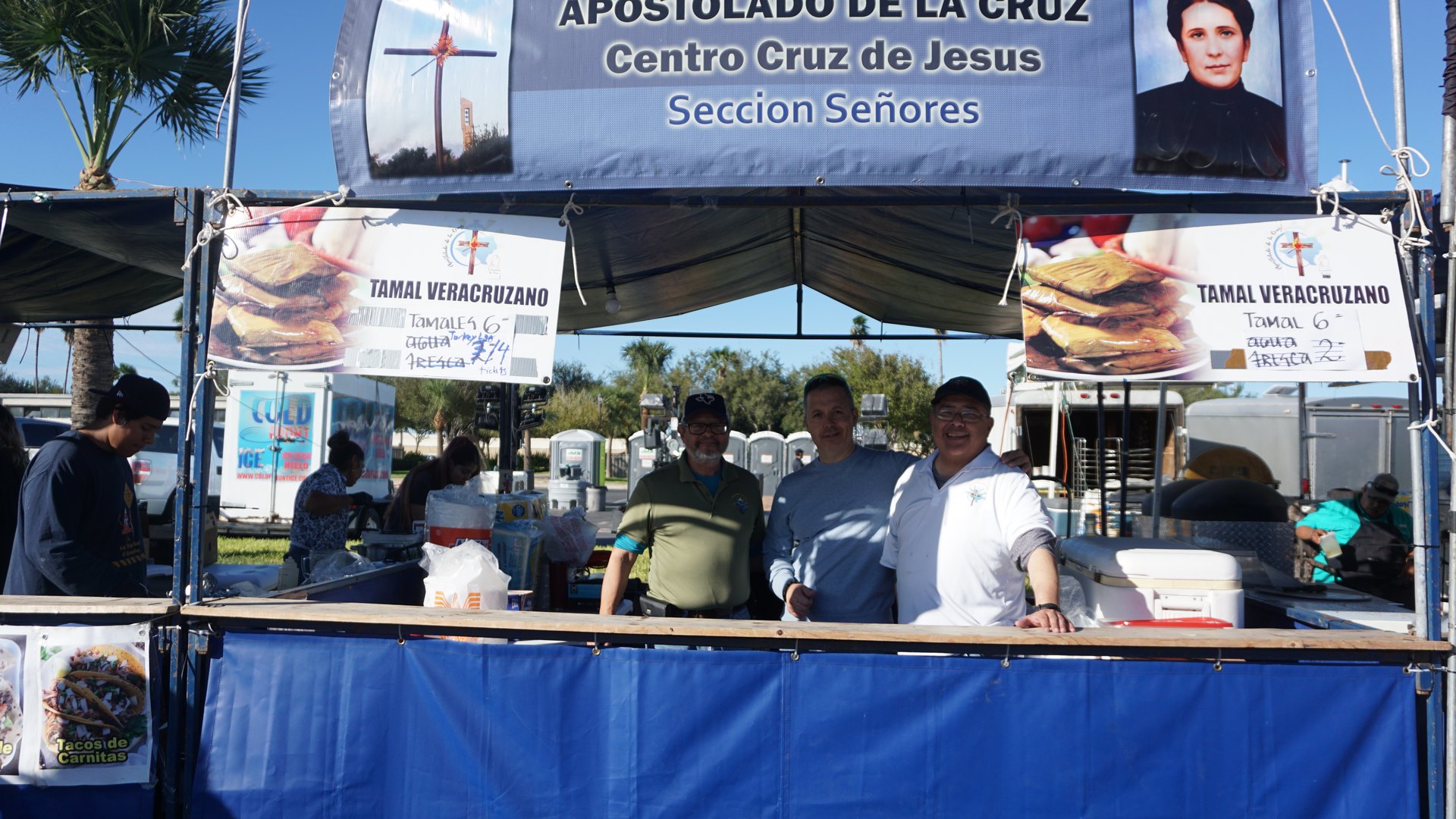 Booth attendants gather behind a tamale counter with food containers and menu signs.