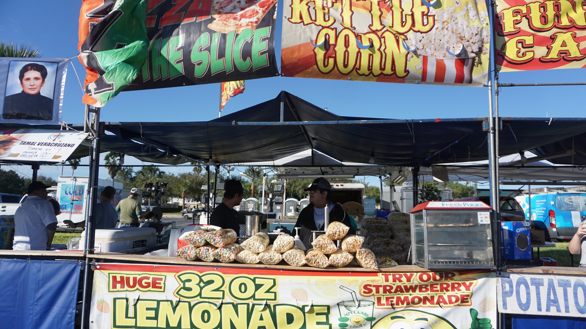 Festival vendors stand behind a snack counter piled with popcorn bags beneath kettle corn and pizza banners.