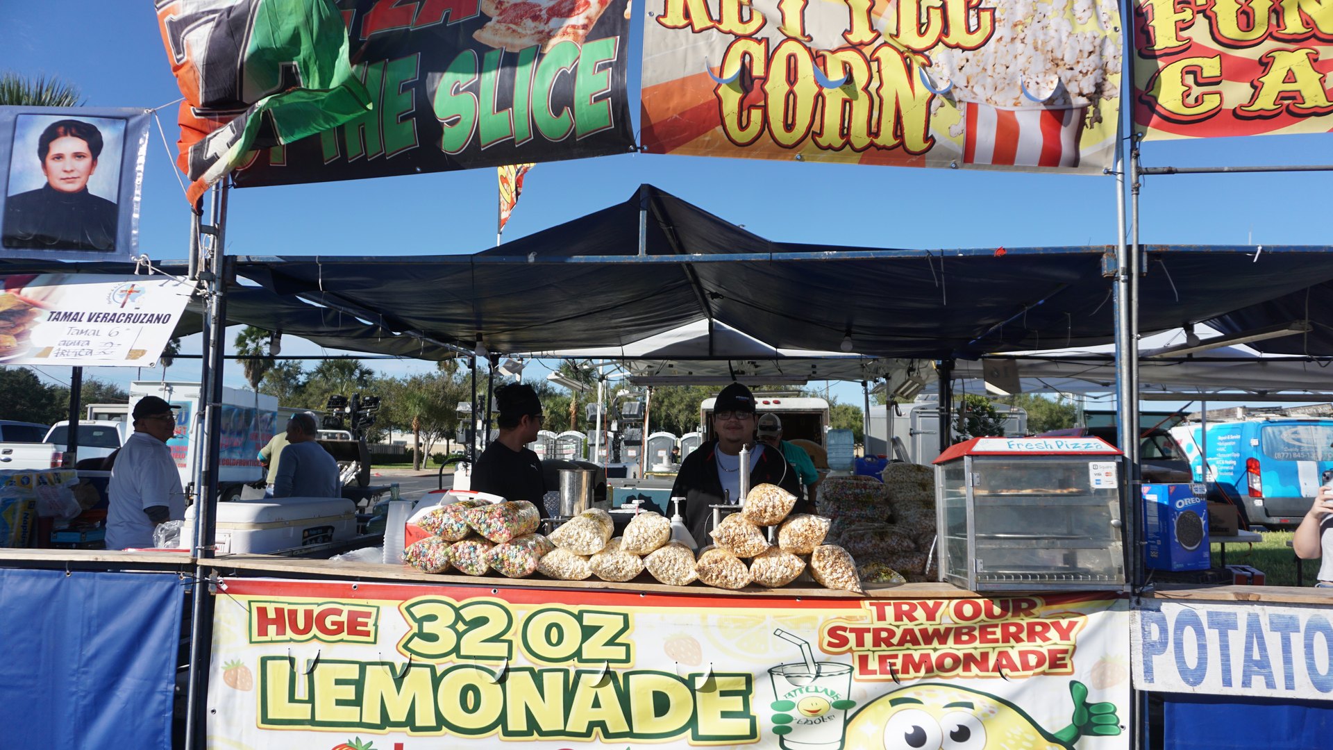 A festival snack stand displays bags of popcorn beneath colorful kettle corn and lemonade signs.