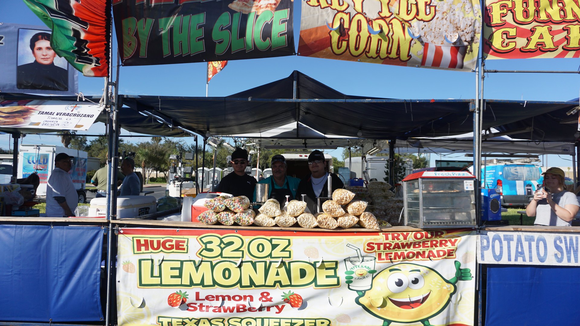 A wide view of a festival snack booth with popcorn bags, lemonade signs and a blue counter.