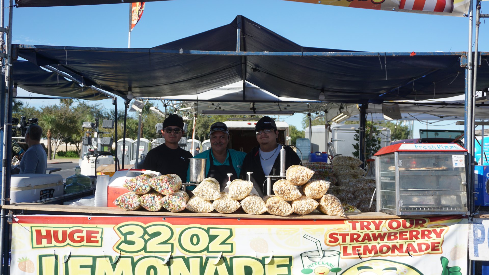 Three snack booth attendants pose behind a display of popcorn bags and a bright lemonade sign.