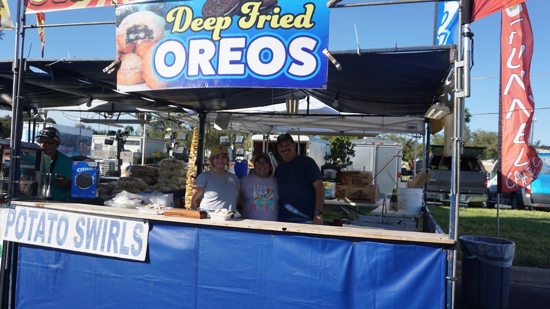 Three people pose at a blue festival booth beneath a Deep Fried Oreos banner and a Potato Swirls sign.