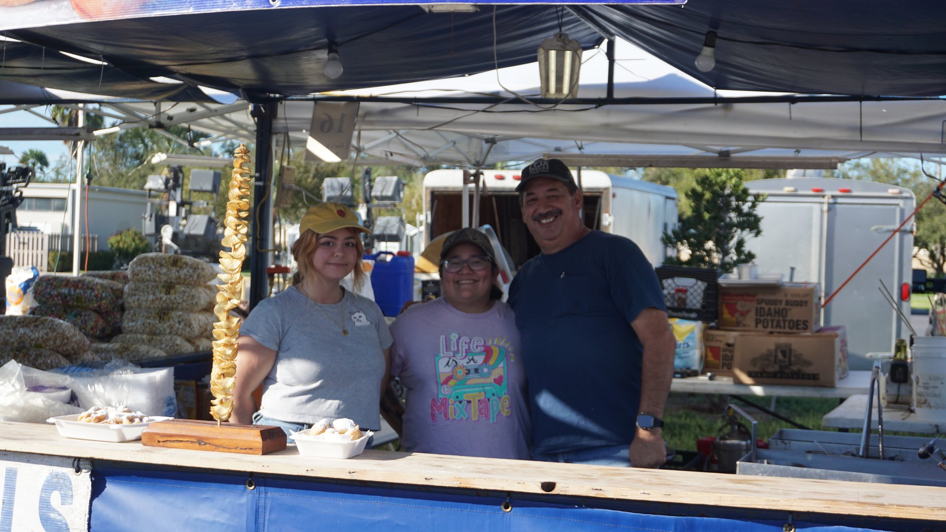 Three smiling snack booth attendants pose behind a counter with cooking supplies and bags of popcorn.