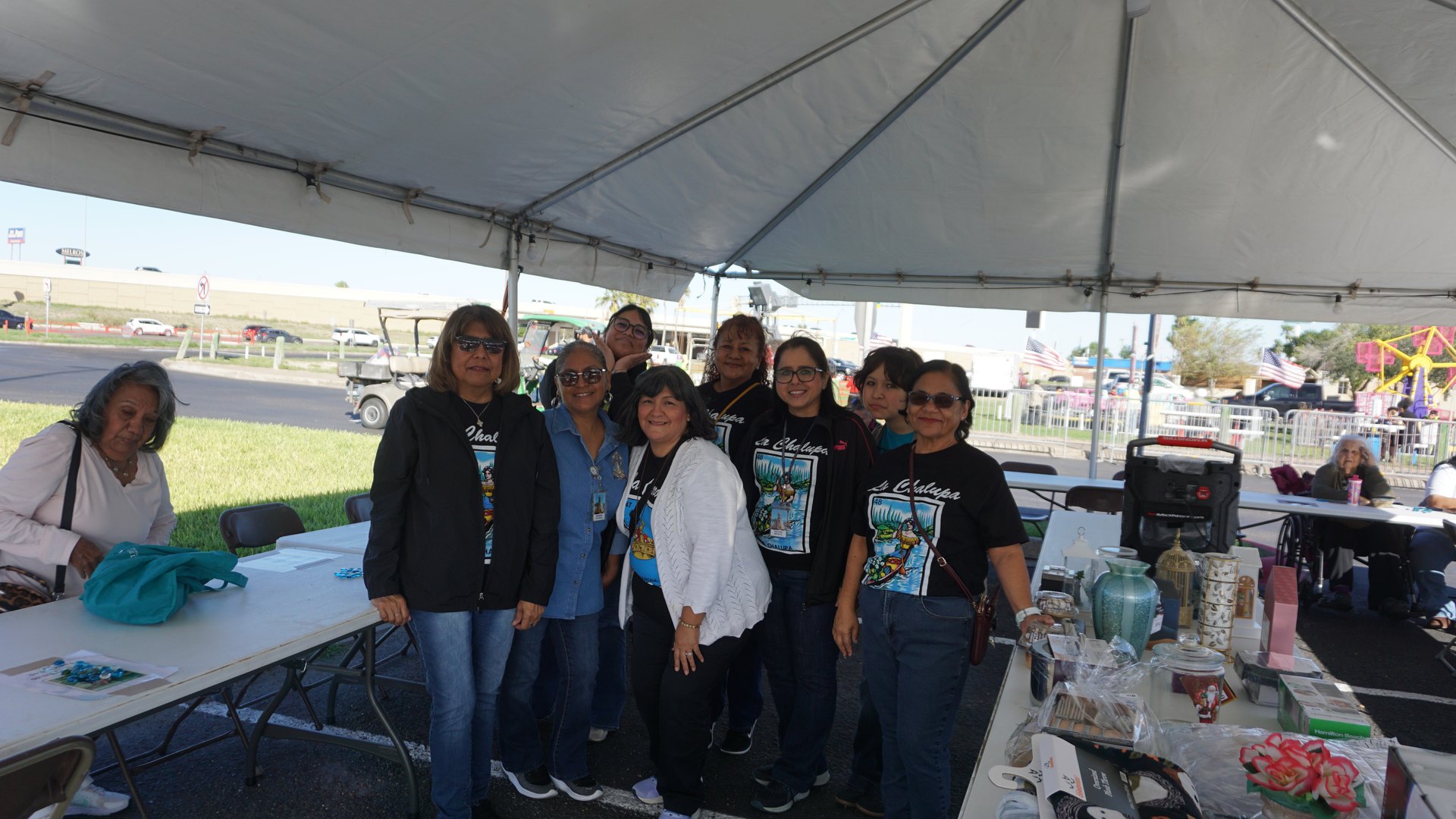 A group poses beneath a white festival tent beside tables displaying decorative items and gifts.
