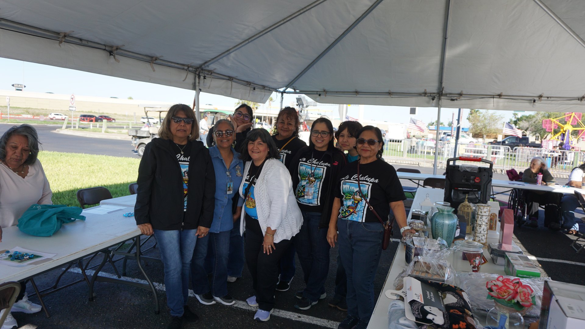 Festival booth participants gather beneath a white tent beside tables of decorative items.