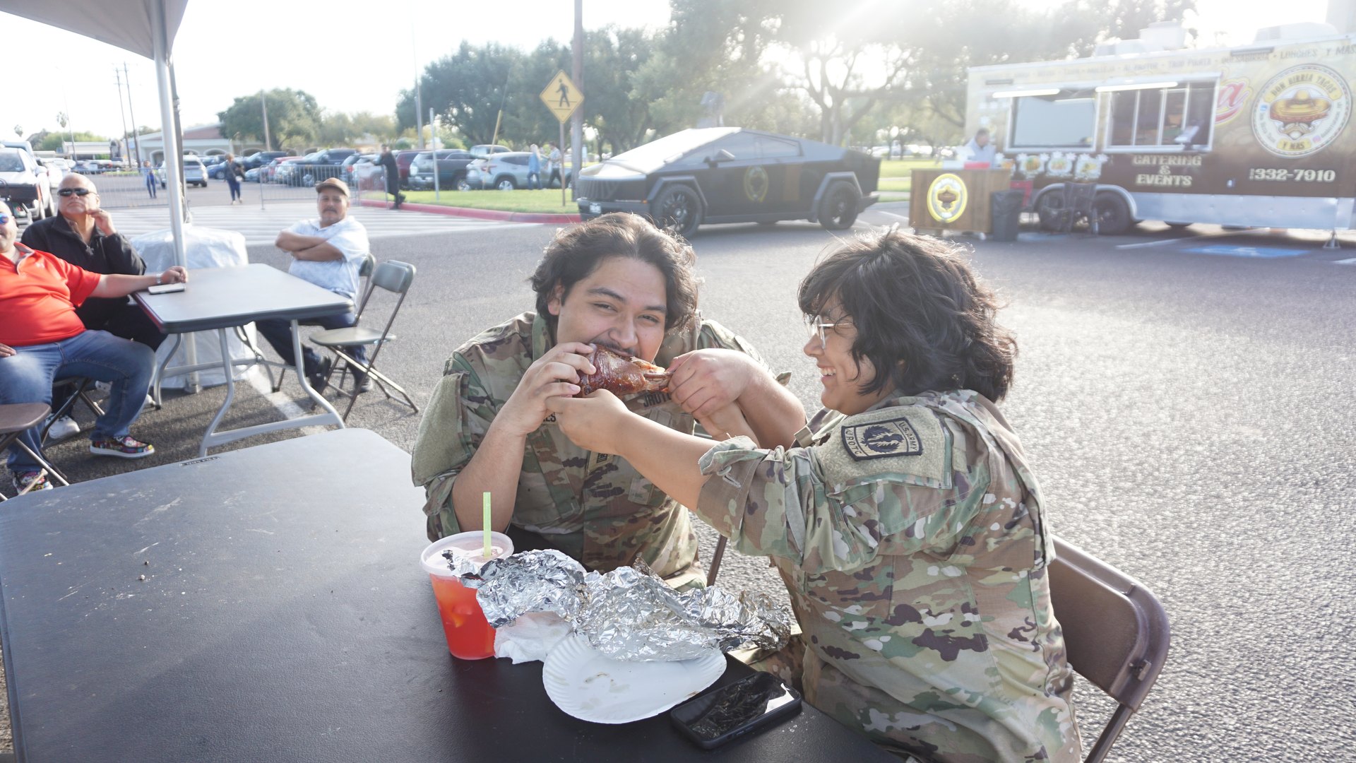 Two people in camouflage clothing enjoy food at a picnic table at the festival.