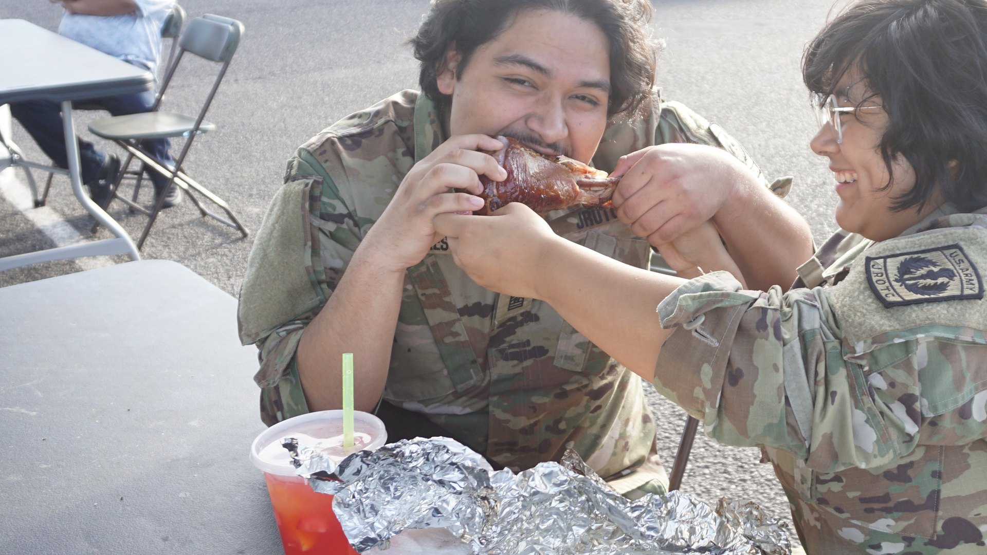 A close view of two people in camouflage clothing sharing a playful moment while eating at a festival table.