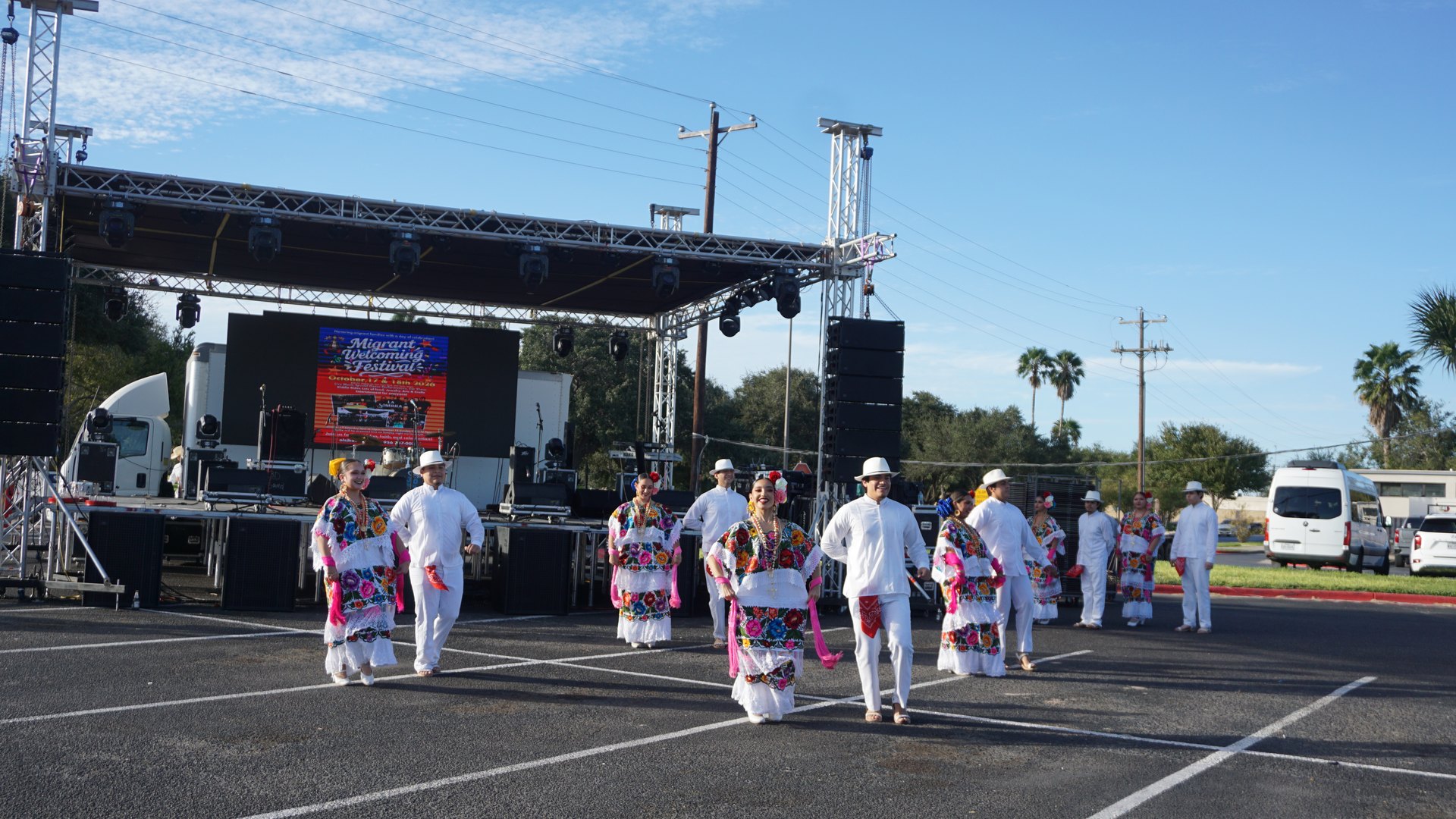 Dancers in floral embroidered dresses and white outfits gather in front of the outdoor festival stage.