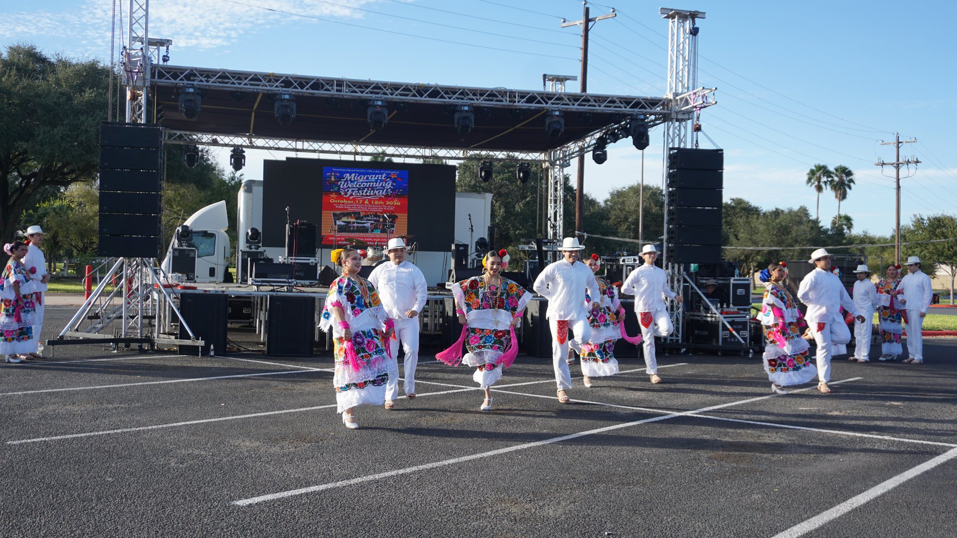 Dancers perform in colorful floral dresses and white outfits with hats in front of the festival stage.