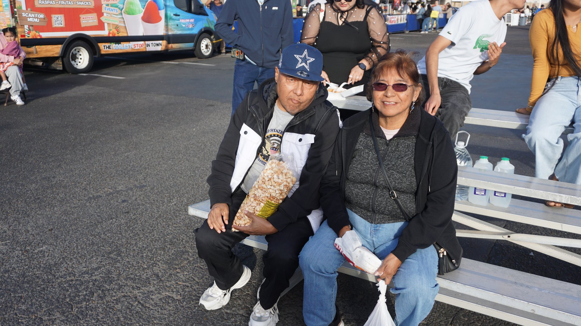 Two festival guests sit together on bleachers with snacks, with food vendors behind them.
