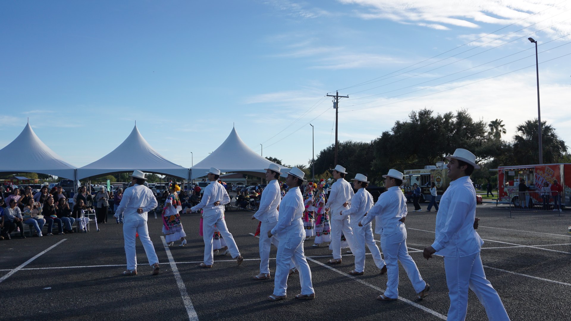 Dancers perform beside festival tents and a seated audience under a blue sky.