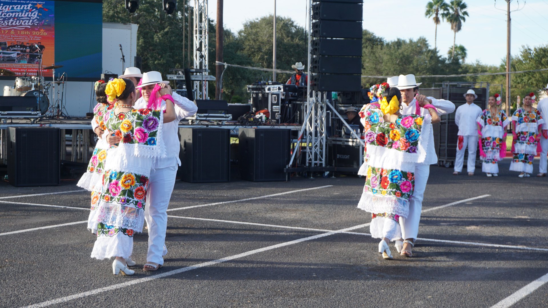 Dance partners in white clothing and colorful floral embroidery turn in front of the stage.