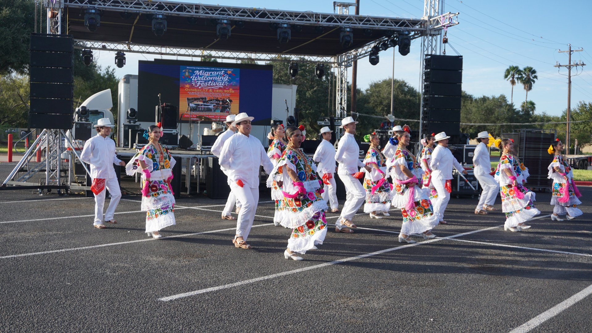 Dancers in colorful embroidered dresses and white outfits perform together beneath the festival banner.