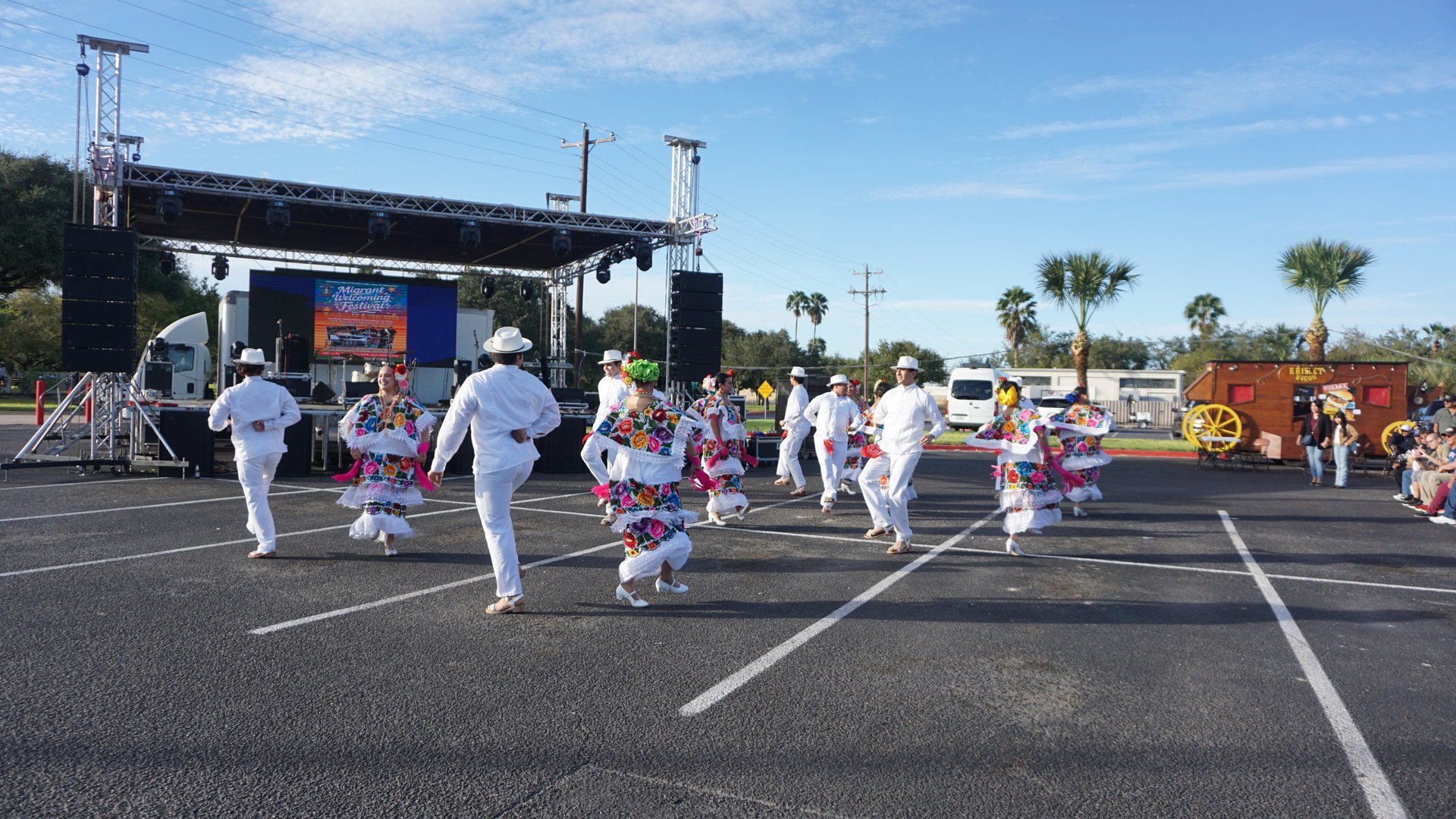 Wide view of the dance group performing in front of the stage beneath a blue sky.