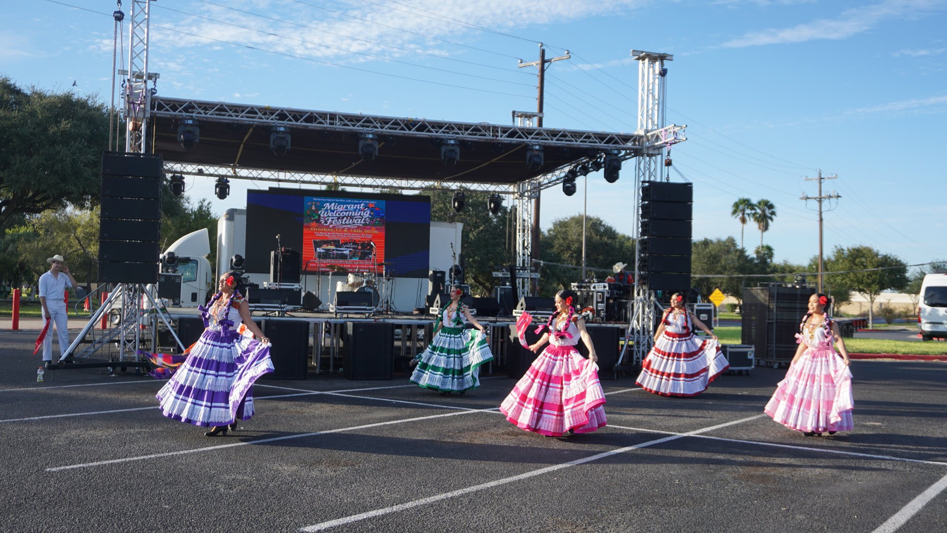 A line of dancers turns in flowing multicolored dresses with the festival stage behind them.