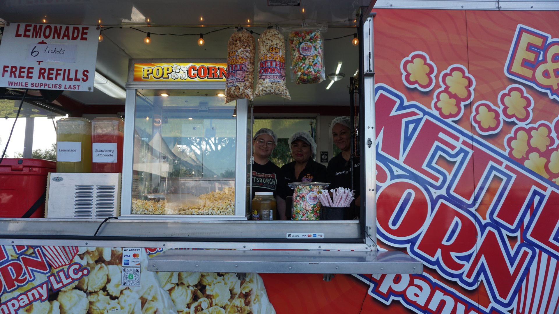 Workers stand at the window of a kettle corn trailer filled with popcorn bags.