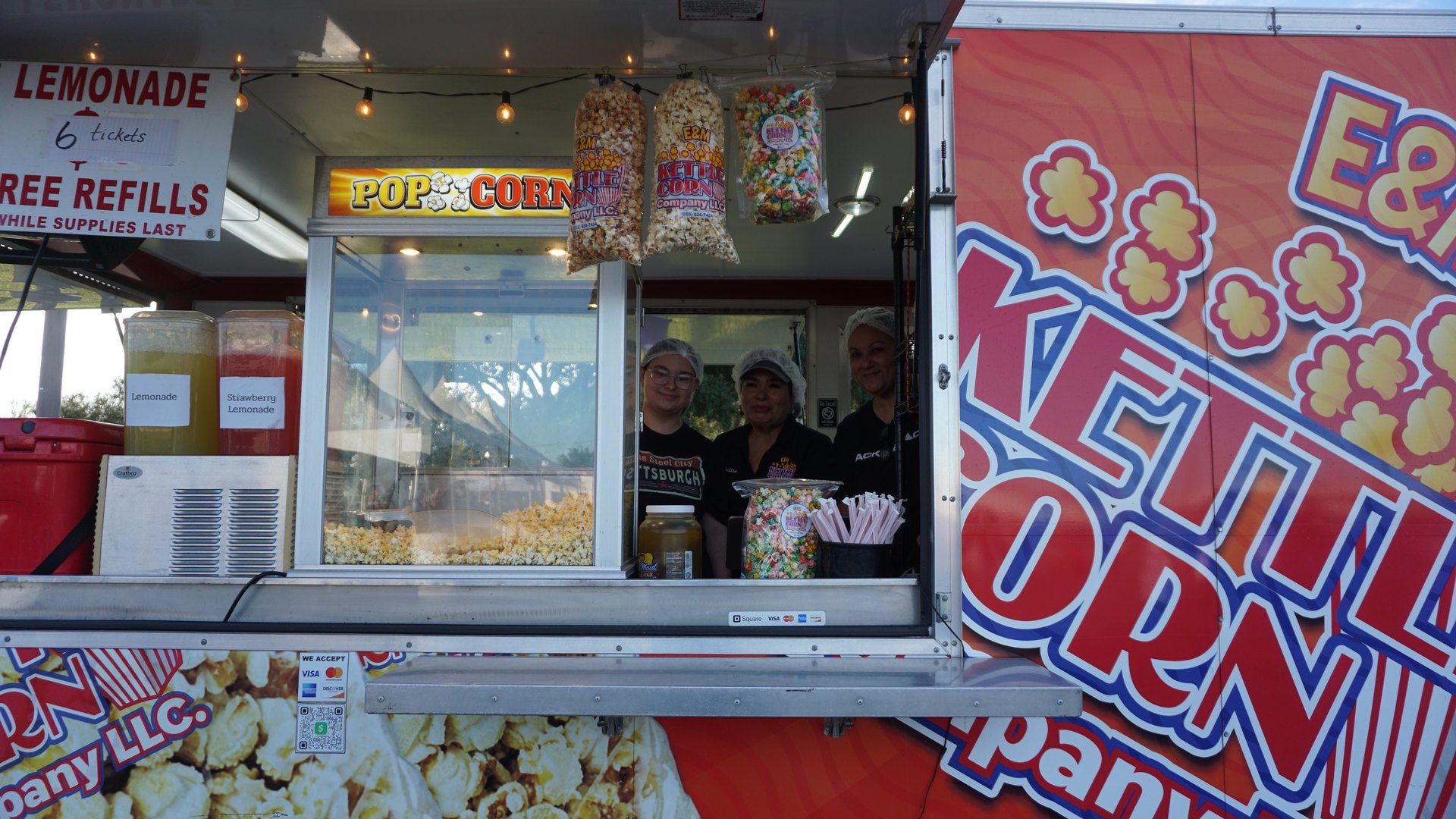 Kettle corn vendors pose behind the service window of their colorful concession trailer.