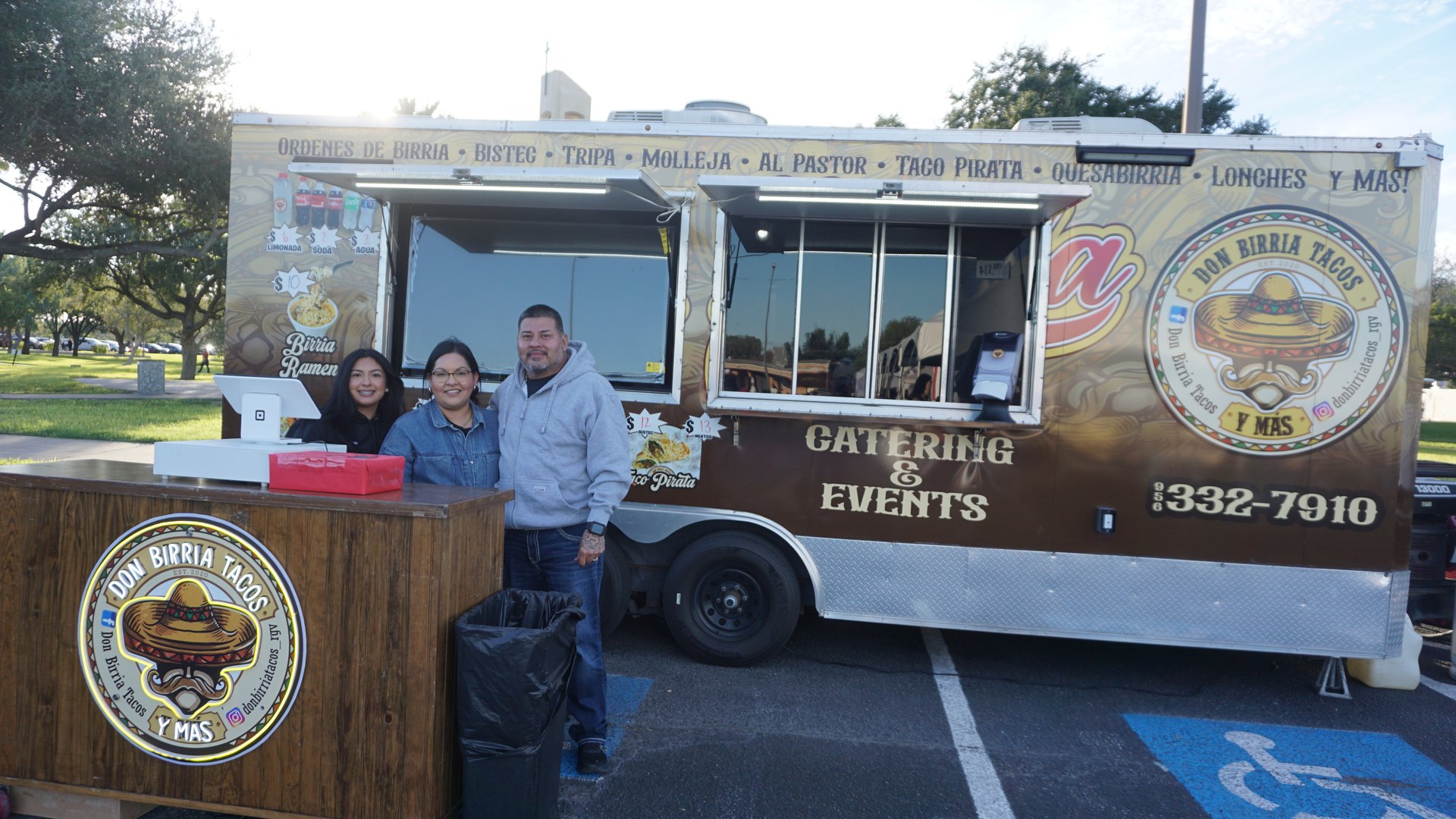 Food vendors stand together at a wooden counter beside their taco trailer.