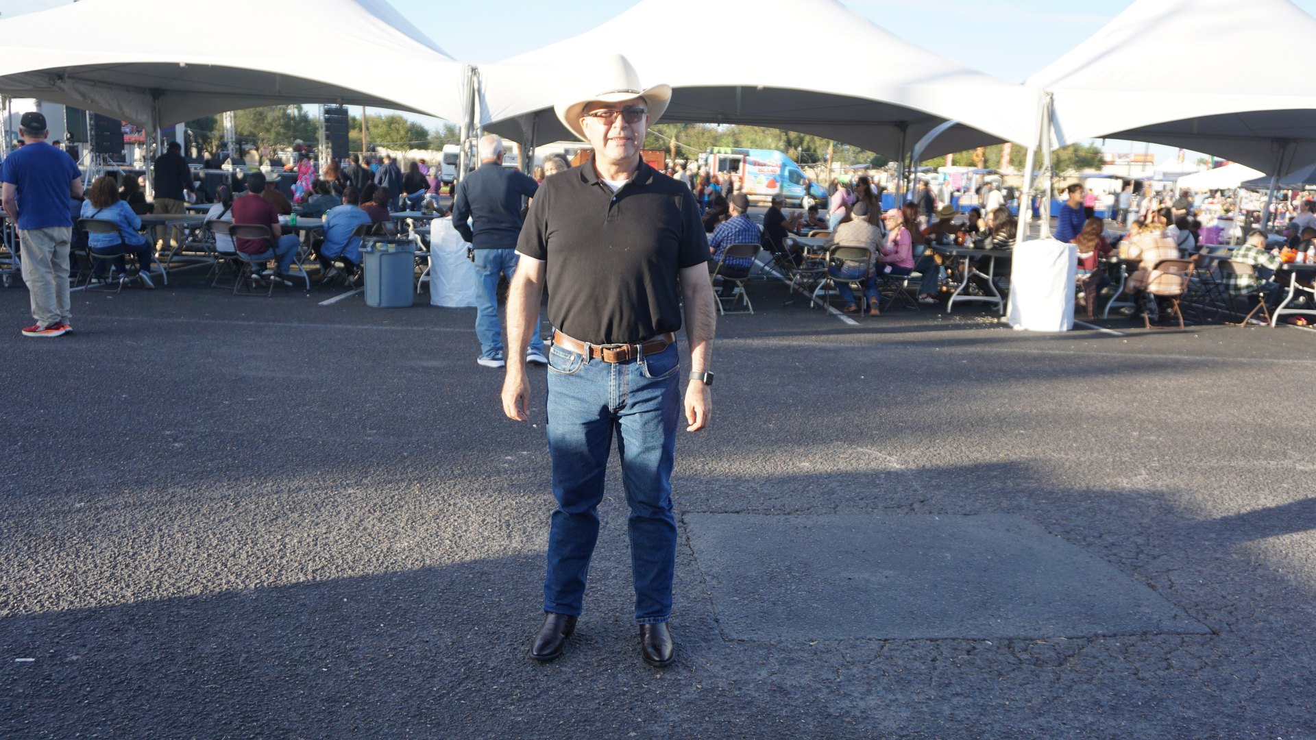 A guest in a cowboy hat and black shirt stands smiling near the white festival tents.