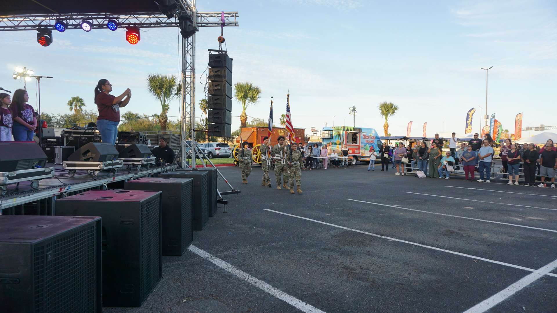 A speaker stands at the edge of the festival stage as uniformed flag bearers approach.