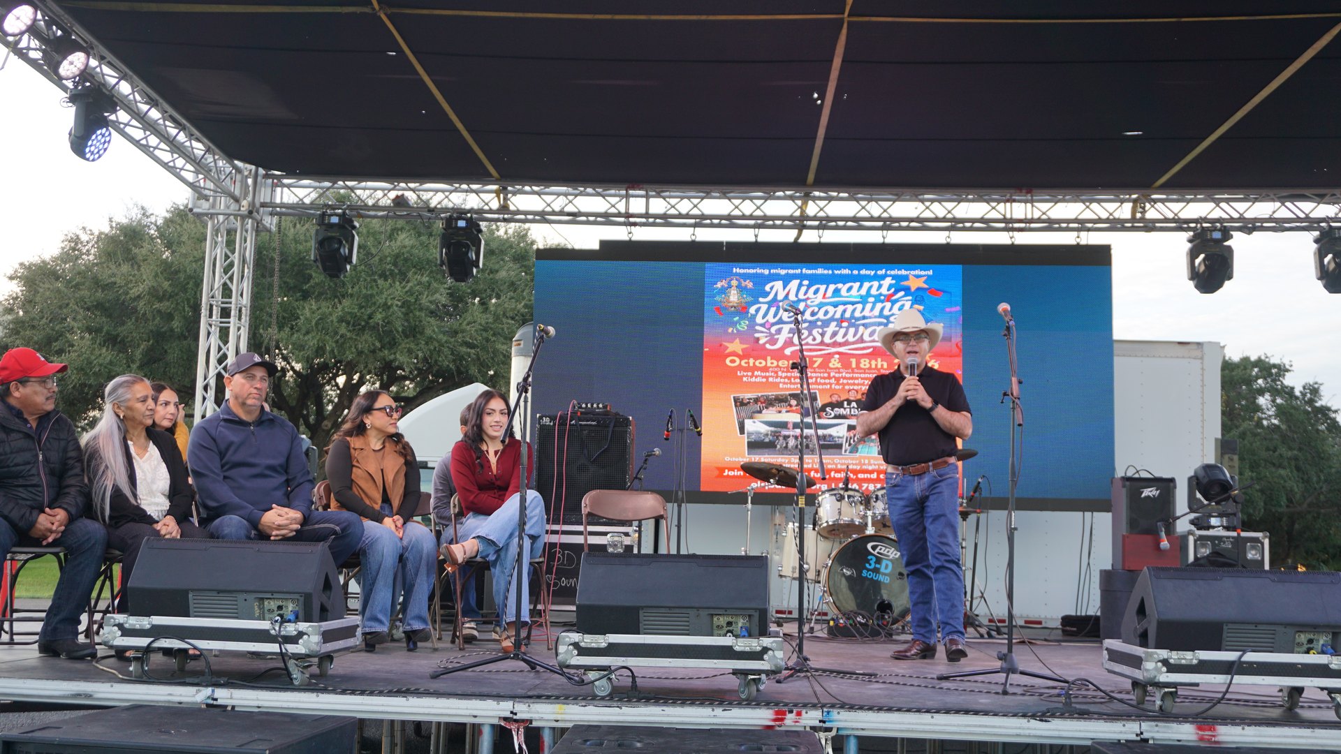 A man in a white cowboy hat speaks into a microphone beside a seated group on the festival stage.