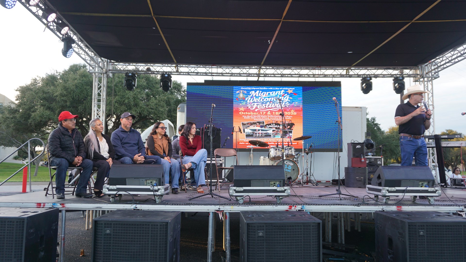A speaker addresses seated guests onstage with the festival sign and musical equipment behind them.