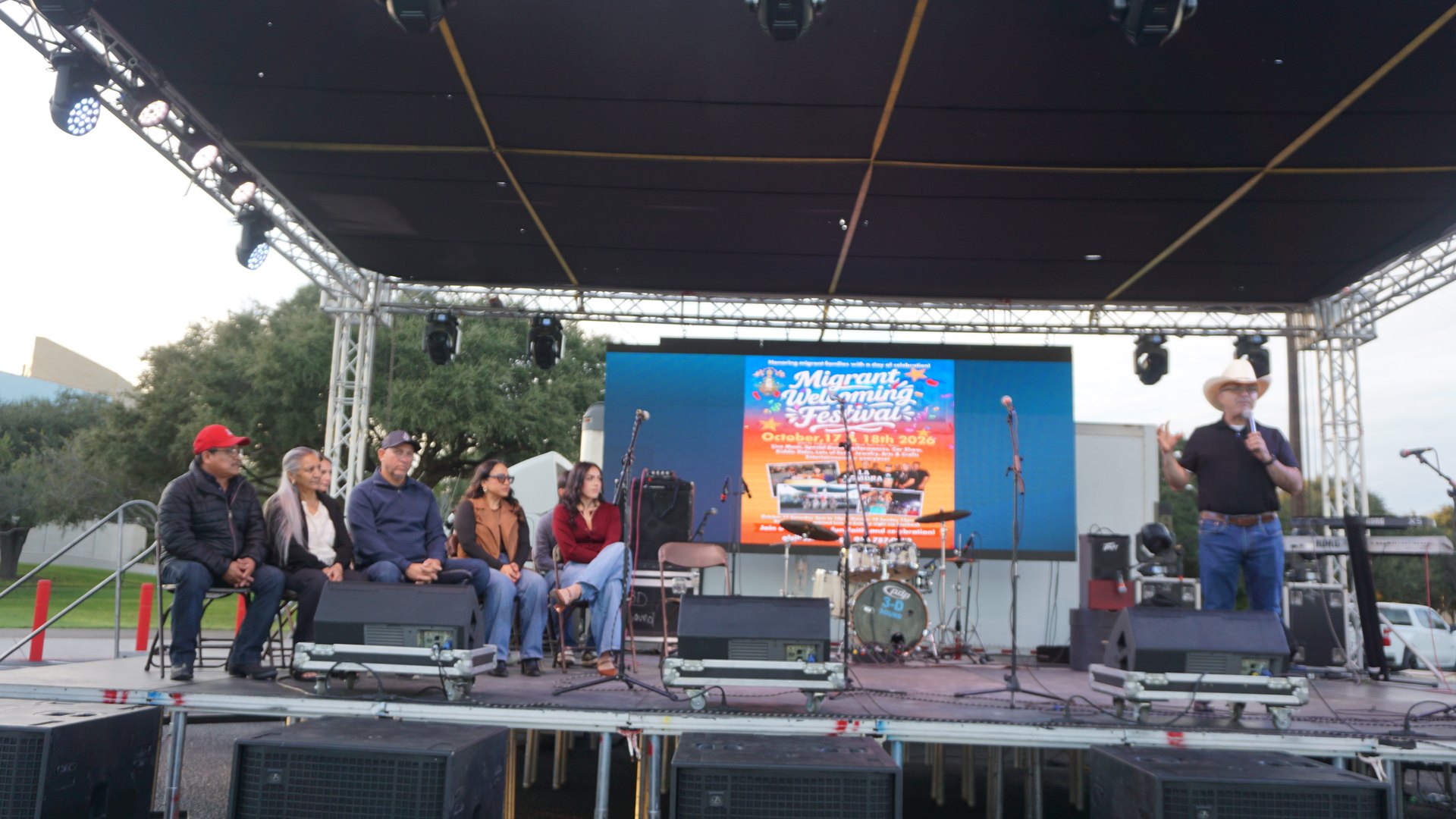 A wide view of a man speaking on the festival stage while several guests sit nearby.