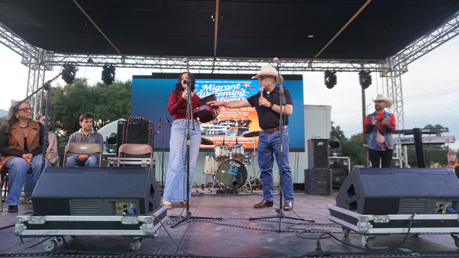 A young woman holding a plaque stands with a microphone beside a man in a white cowboy hat onstage.