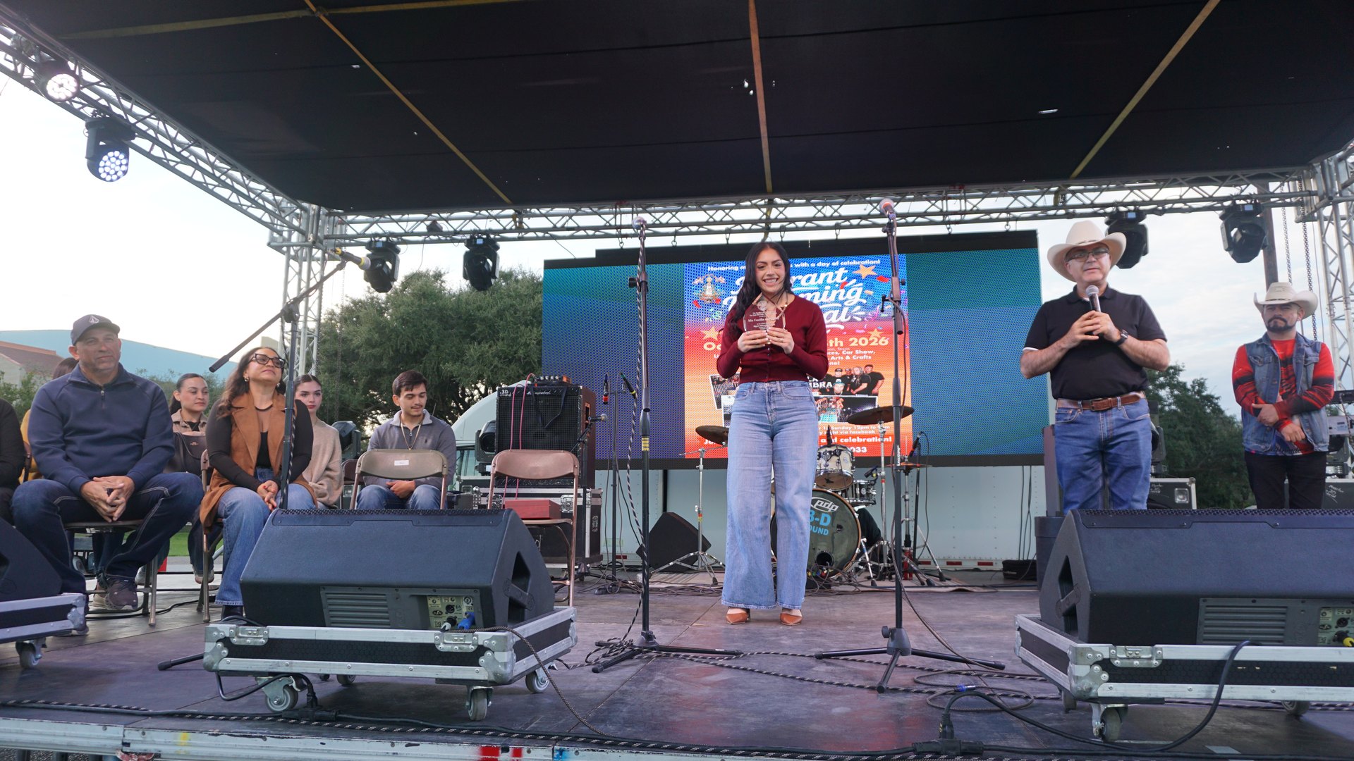 Two speakers stand at microphones before the festival sign with guests seated to the side.