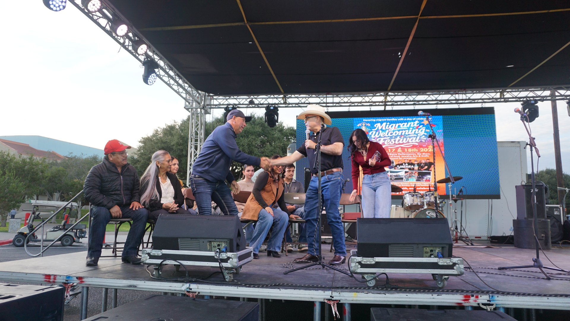 A man in a white cowboy hat reaches toward a seated guest during the onstage presentation.