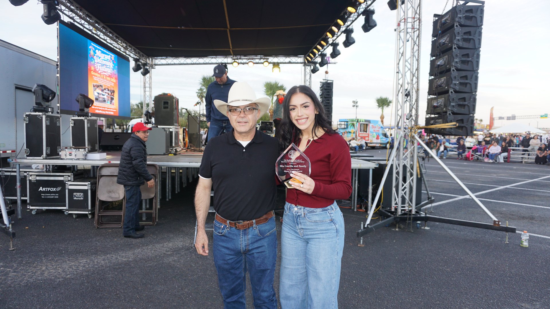 A man in a white cowboy hat and a smiling young woman holding a plaque pose beside the festival stage.