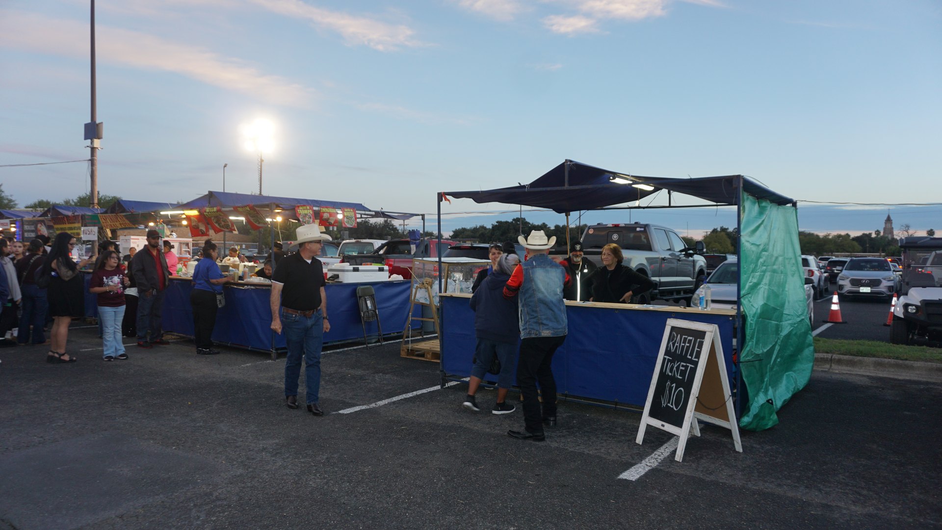 A wide evening view of festival booths, visitors, and food vendors under the open sky.