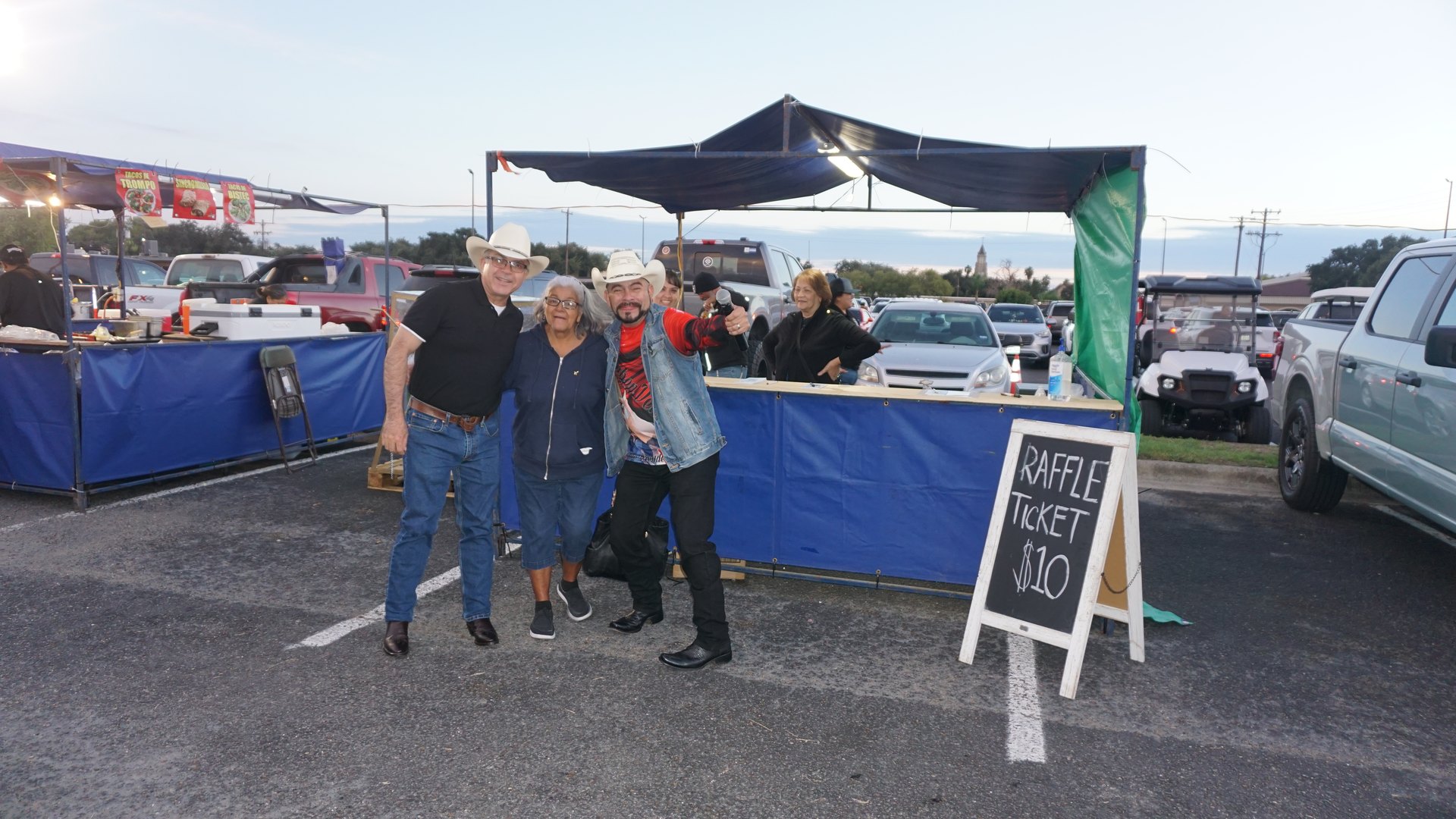 Three festival guests pose together beside a blue booth near the white tents.