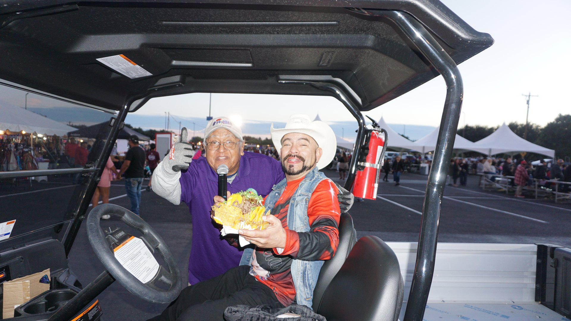 Two men pose in a utility vehicle, one holding a microphone and the other a plate of food, at the festival.